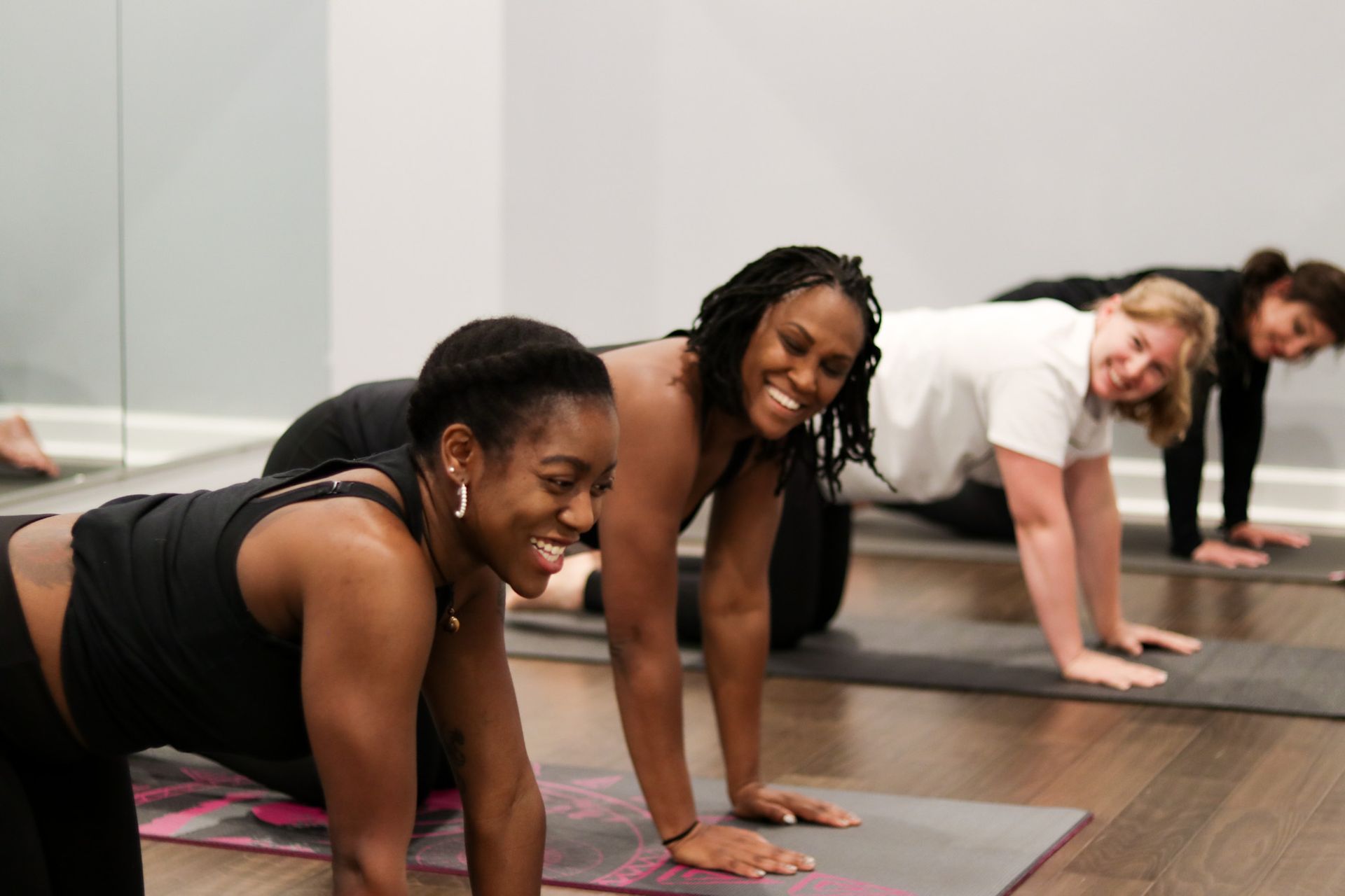 Six women seated in a yoga class, in a brightly lit studio, on colorful mats, cross-legged.