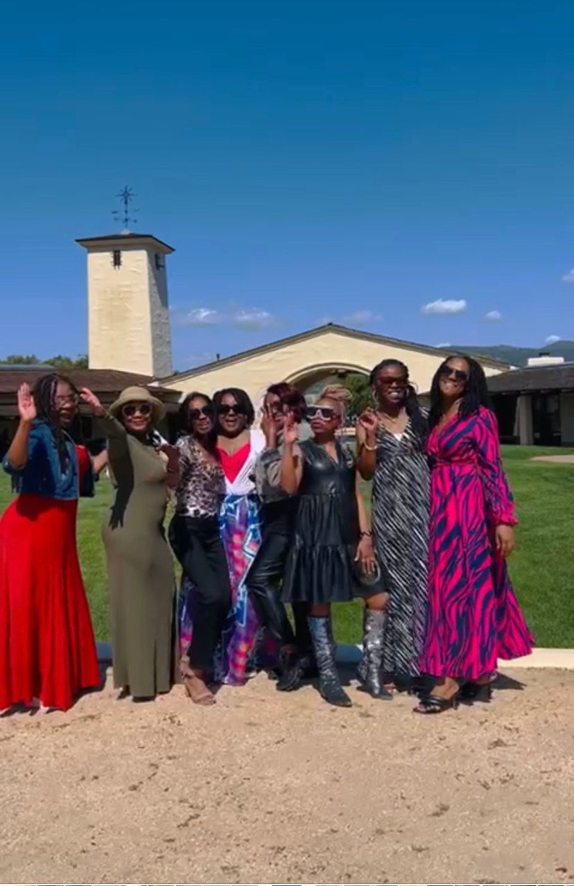 Nine people stand in a line posing outdoors on a sunny day in front of a building with a tower, many waving or smiling.