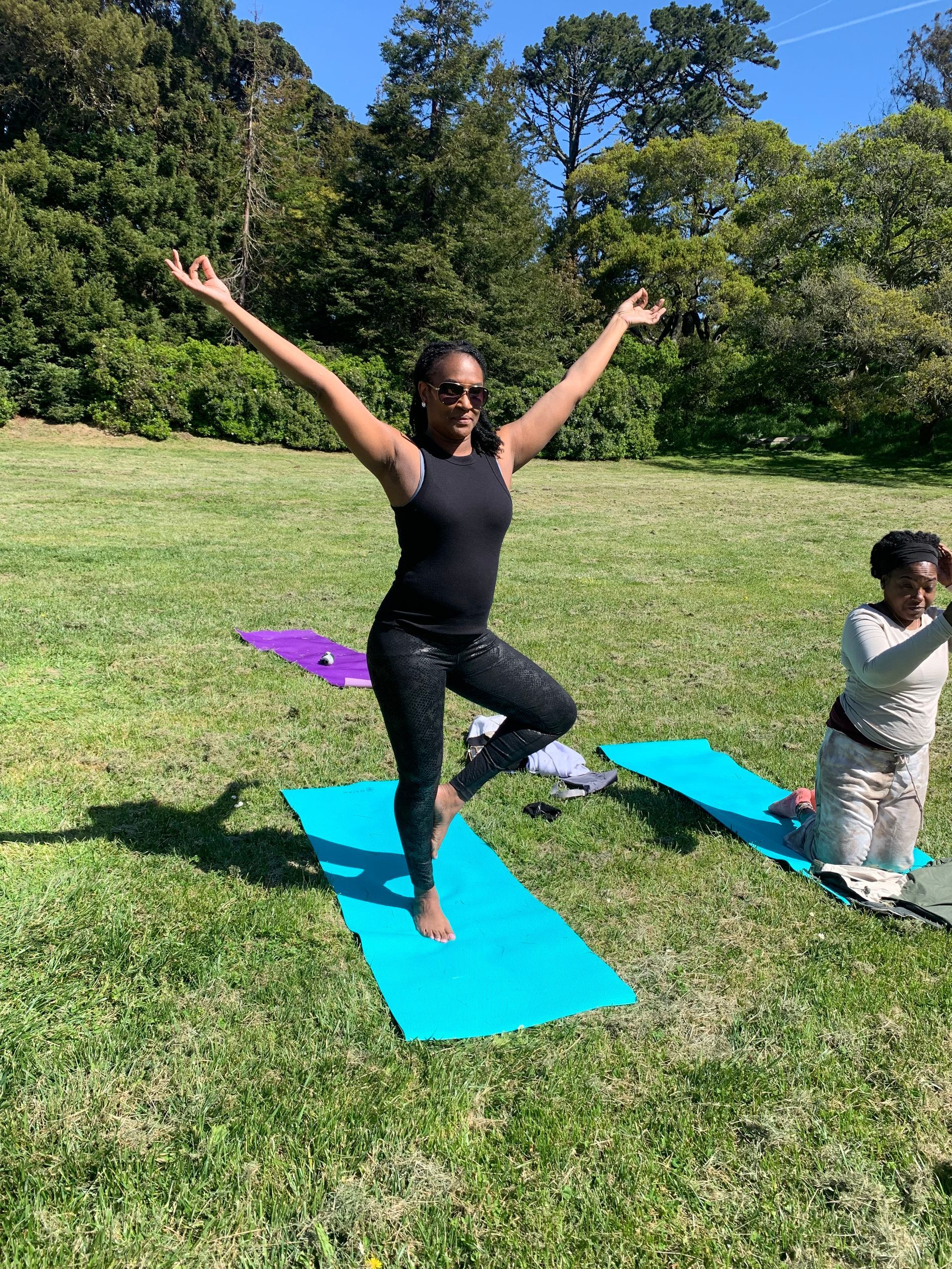 Two people doing yoga in a sunny park; one balances on one leg with arms raised, another kneels on a bright blue mat.