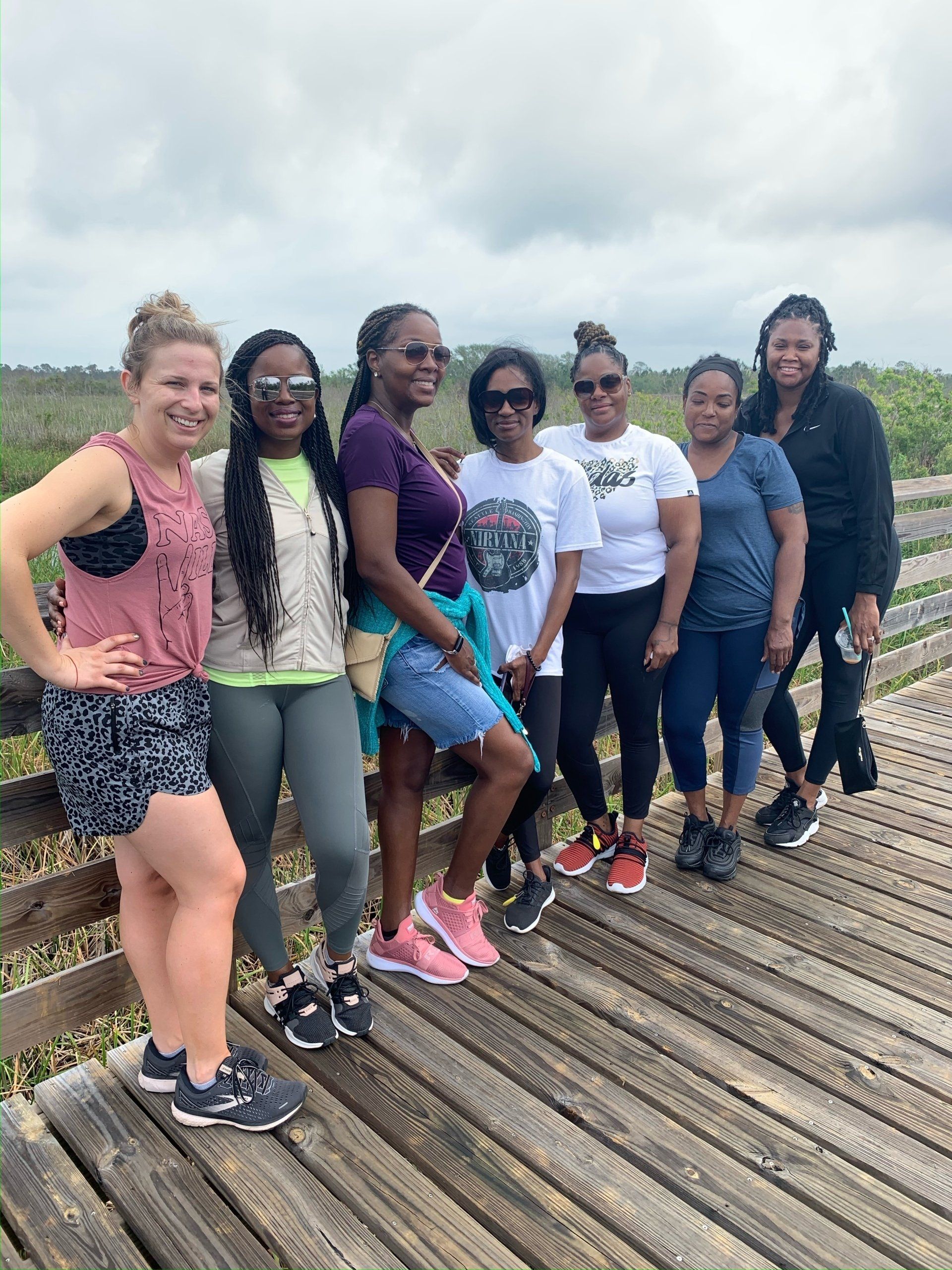 Seven friends stand on a wooden boardwalk outdoors, smiling and posing for a group photo against a natural landscape.