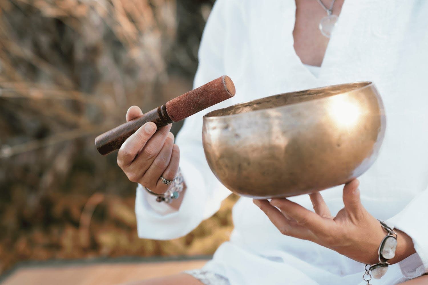 A person in a white shirt holds a bronze singing bowl and a wooden mallet, preparing to play it.