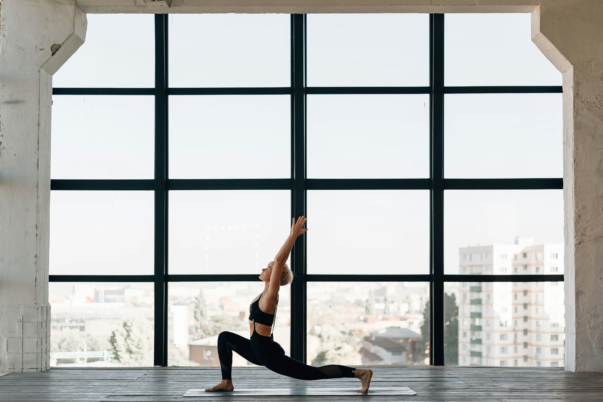 A person in black athletic wear performs a yoga lunge with arms raised against a large window overlooking a city.