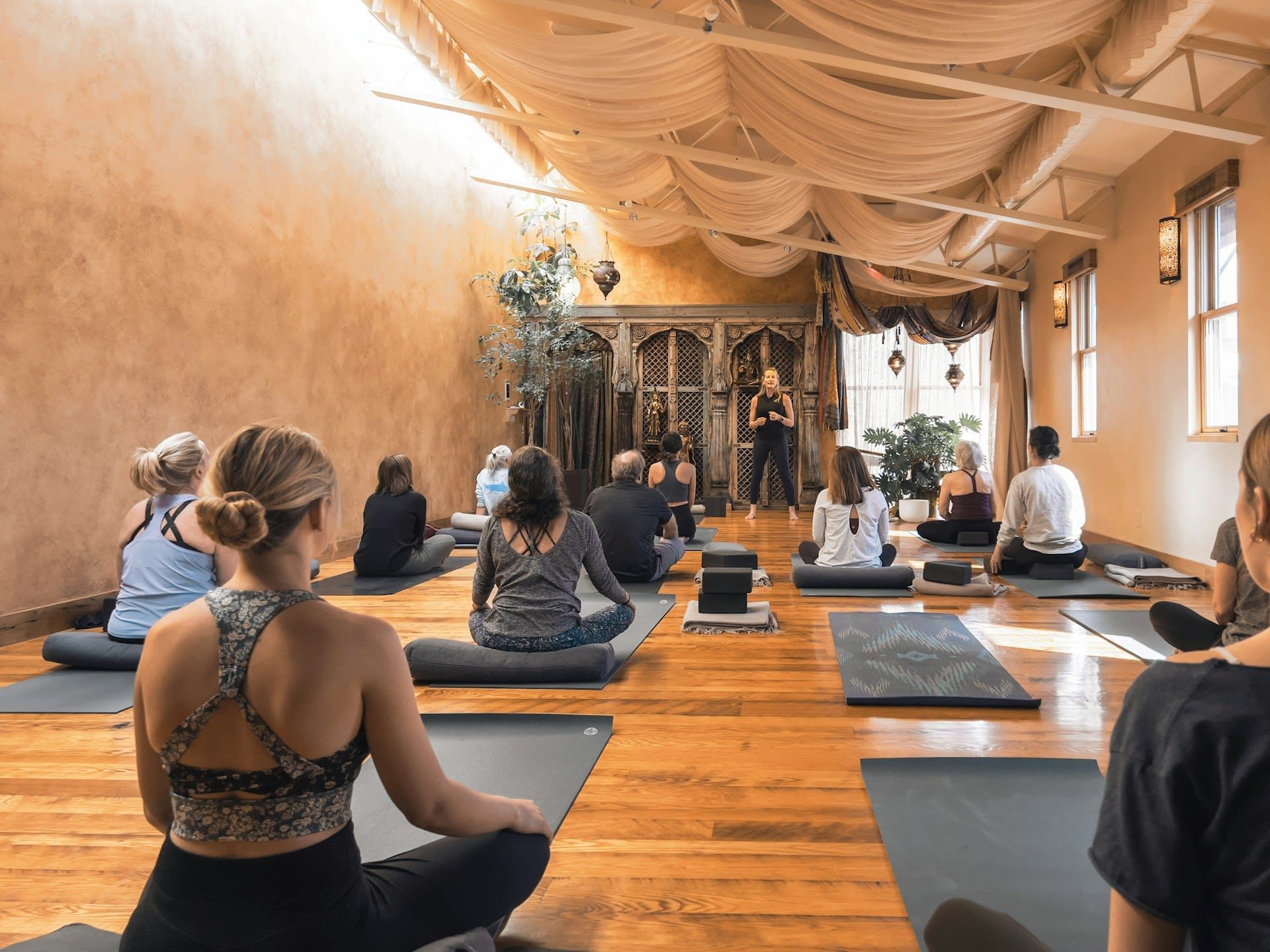 A group sits on mats in a warm-toned yoga studio with draped ceiling fabric, facing an instructor in the distance.