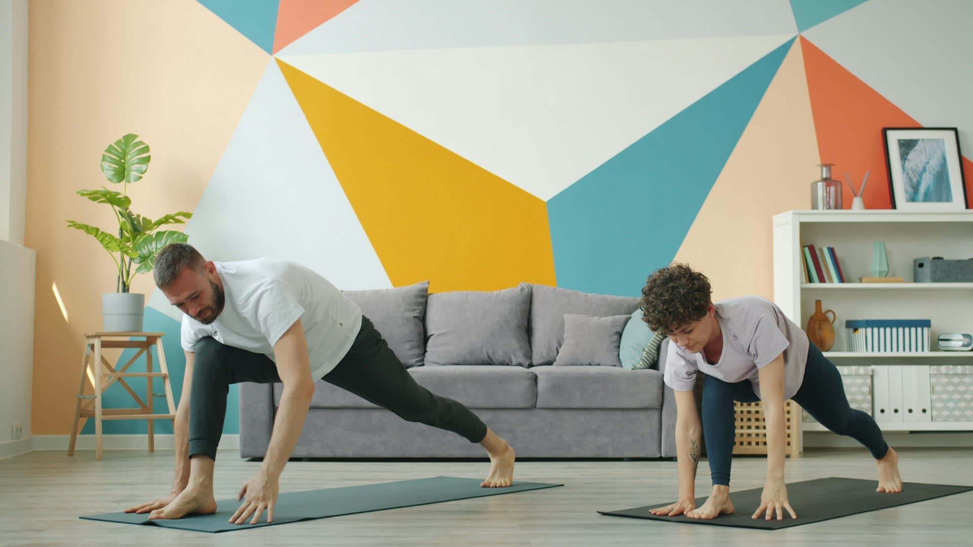 Two people perform a low lunge on yoga mats in a room with a colorful geometric wall, a grey sofa, and a bookshelf.