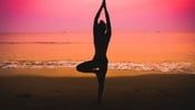 A silhouetted person performs a tree pose on a beach at sunset with a pink and orange sky.