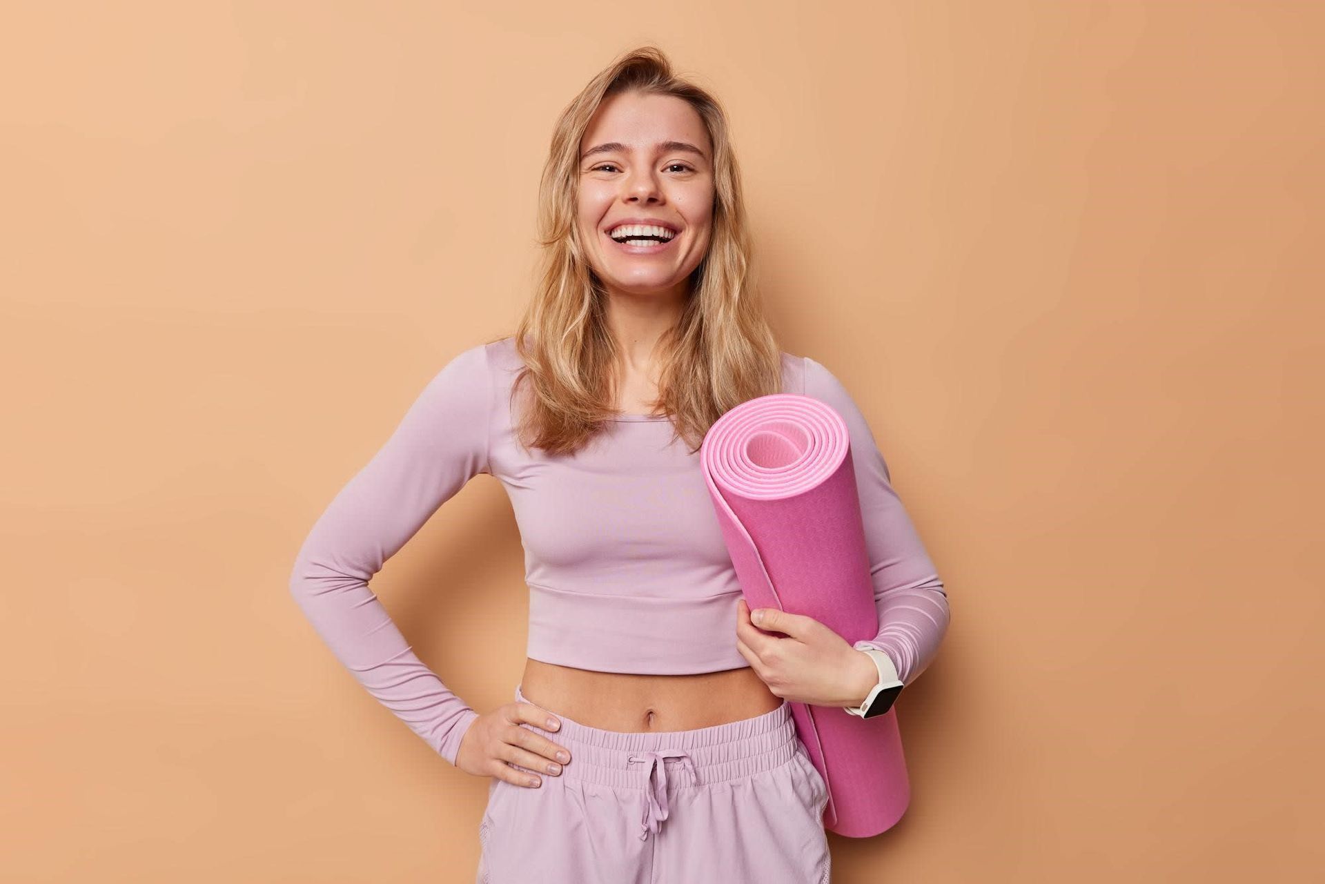 A smiling person wearing lavender activewear stands against a tan background, holding a rolled-up pink yoga mat.