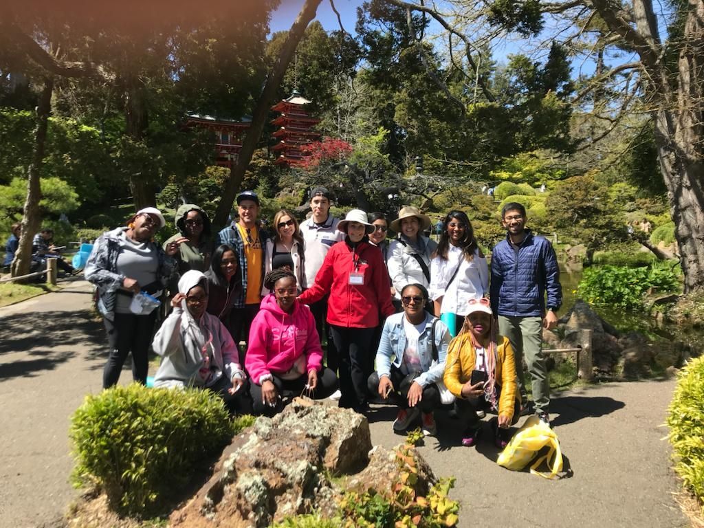 A group of people poses for a photo in a sunny, tree-lined garden with a Japanese-style pagoda in the background.