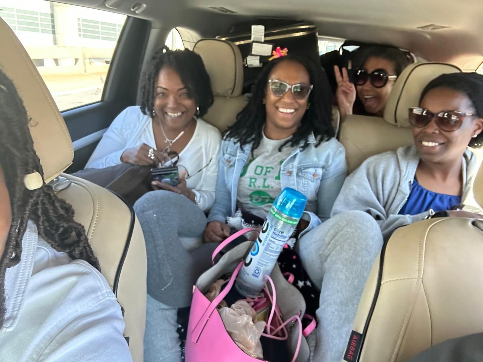 Four people smiling inside a car during a road trip, with a bright pink bag and drinks visible in the foreground.