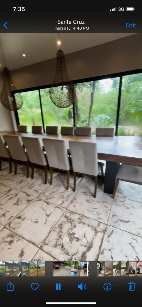 A long wooden dining table with beige chairs faces a large window overlooking a lush green outdoor area.