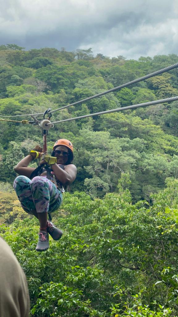 A person wearing a helmet and gloves ziplines through a lush, green forest under a cloudy sky.