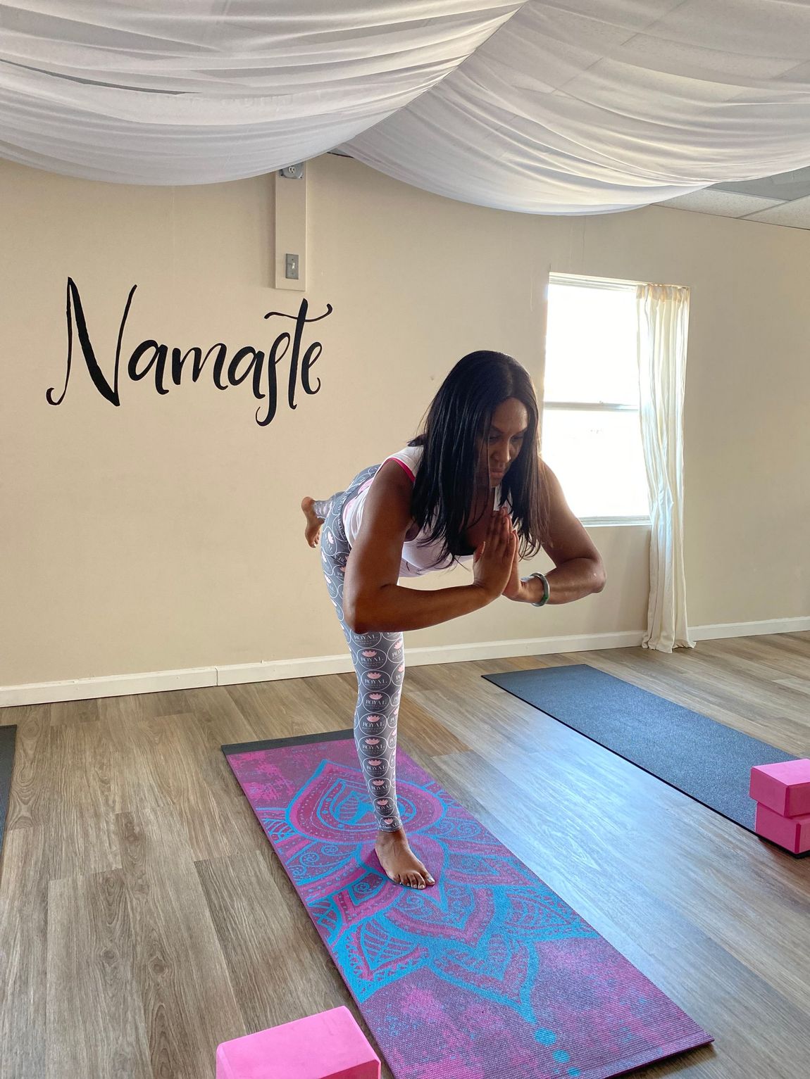 A person practicing Warrior III yoga pose on a patterned blue and purple mat in a studio with 