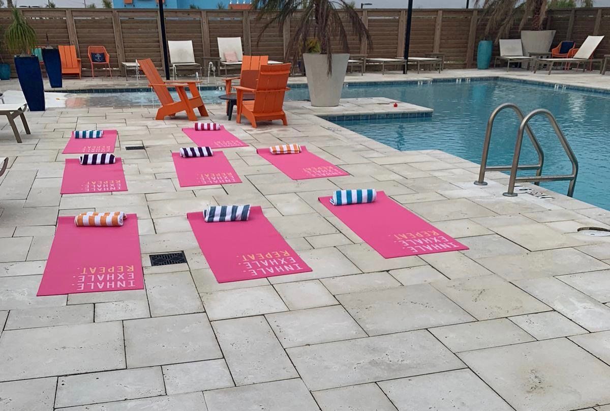 Pink yoga mats with rolled towels are set up on a stone pool deck for an outdoor yoga class by a swimming pool.