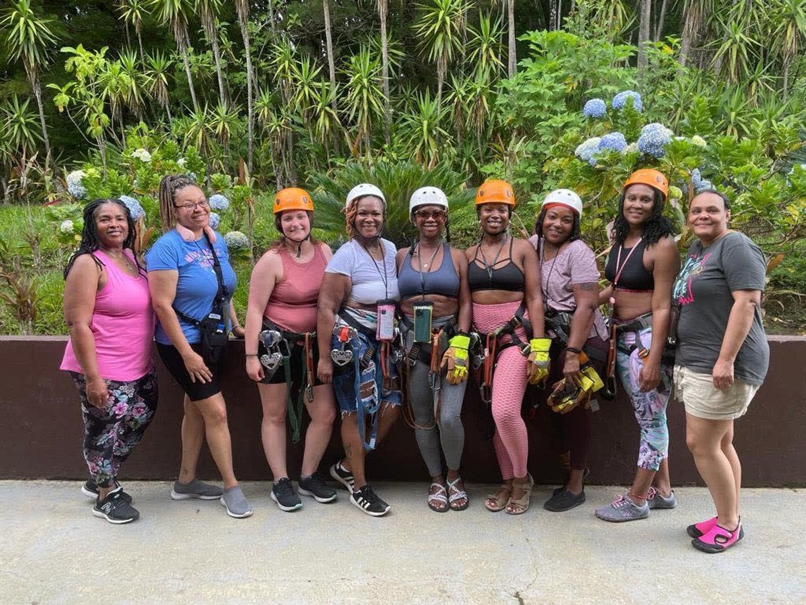 A group of nine people wearing climbing helmets and harnesses pose together in front of a lush, green outdoor setting.