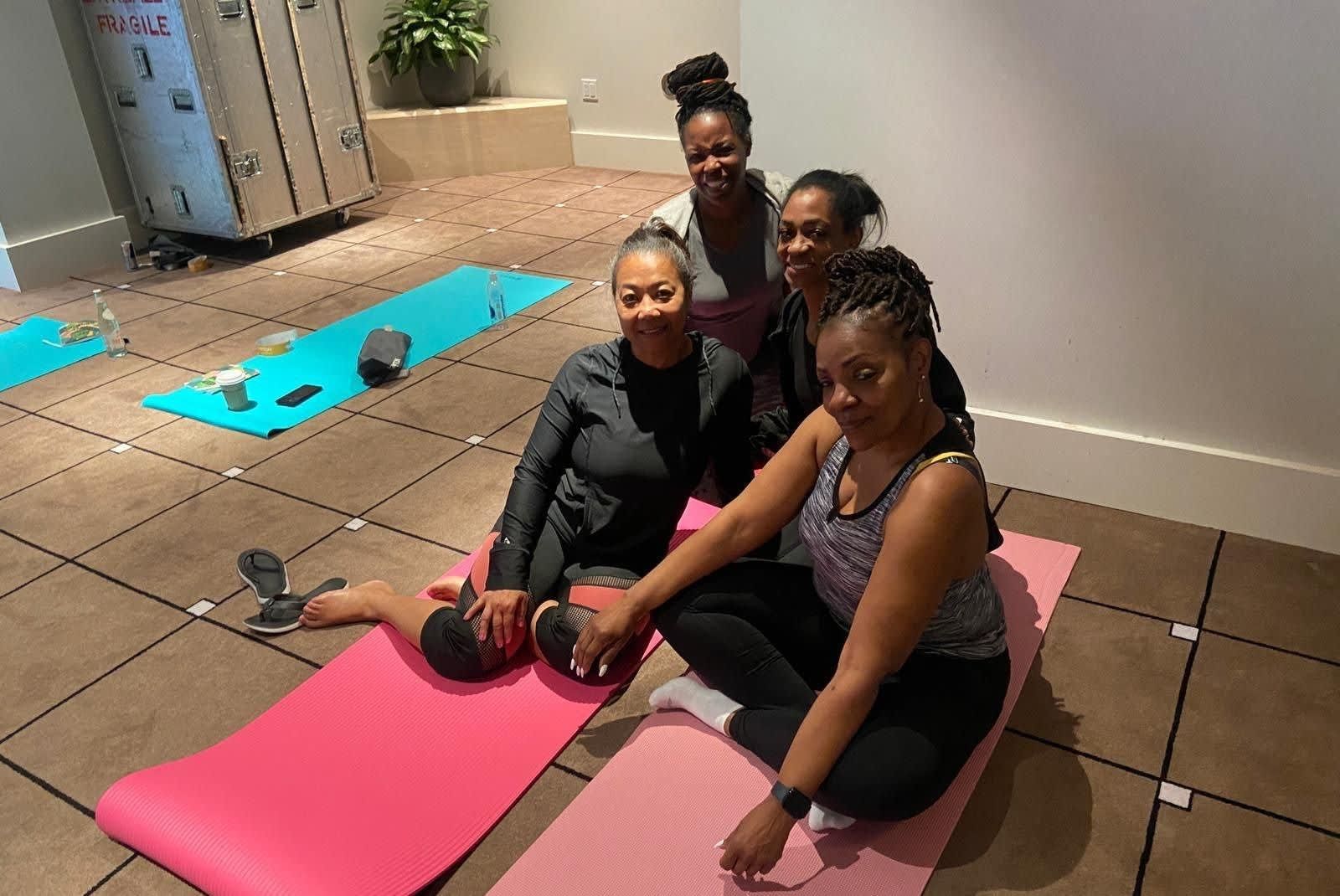 Four people sitting on colorful yoga mats on a tiled floor in a studio space, smiling at the camera.