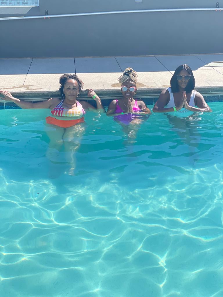 Three people pose in a swimming pool on a sunny day, with arms stretched out or hands together in a gesture.