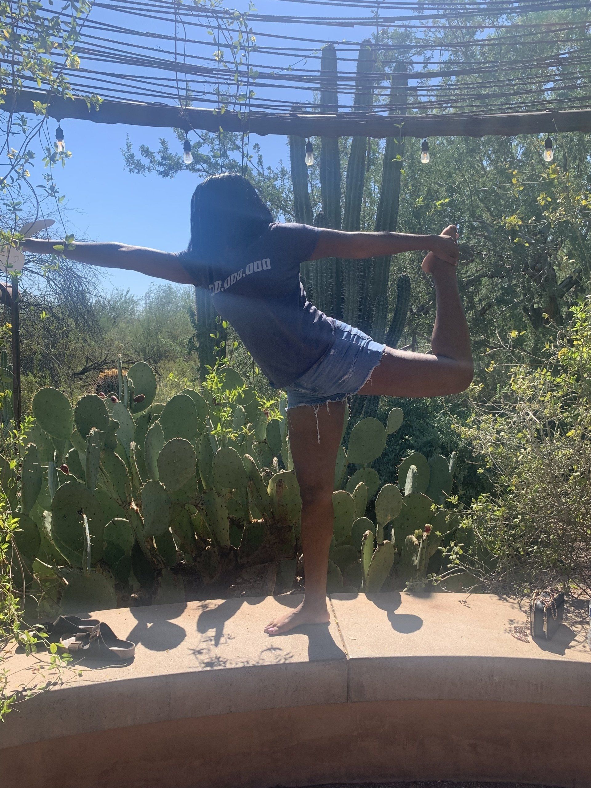 A person in a dark t-shirt and shorts balances on one leg in a dancer yoga pose outdoors near prickly pear cacti.