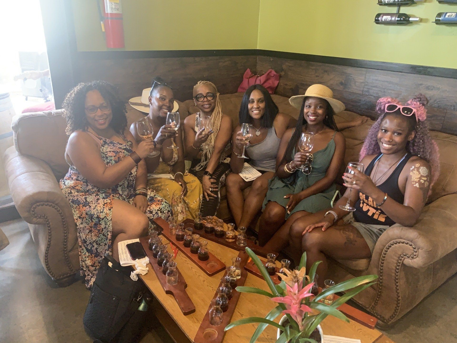 Six people sit together on a large couch at a wine tasting, holding glasses and smiling at the camera.