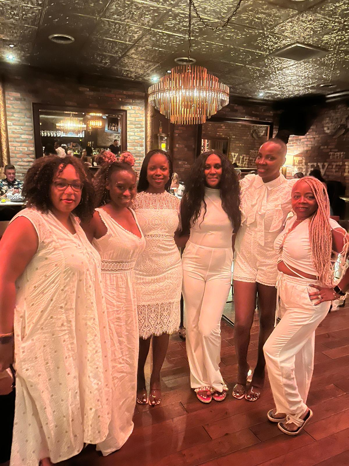 Six individuals in all-white outfits smile for a group photo in a dimly lit, rustic restaurant with a gold chandelier.