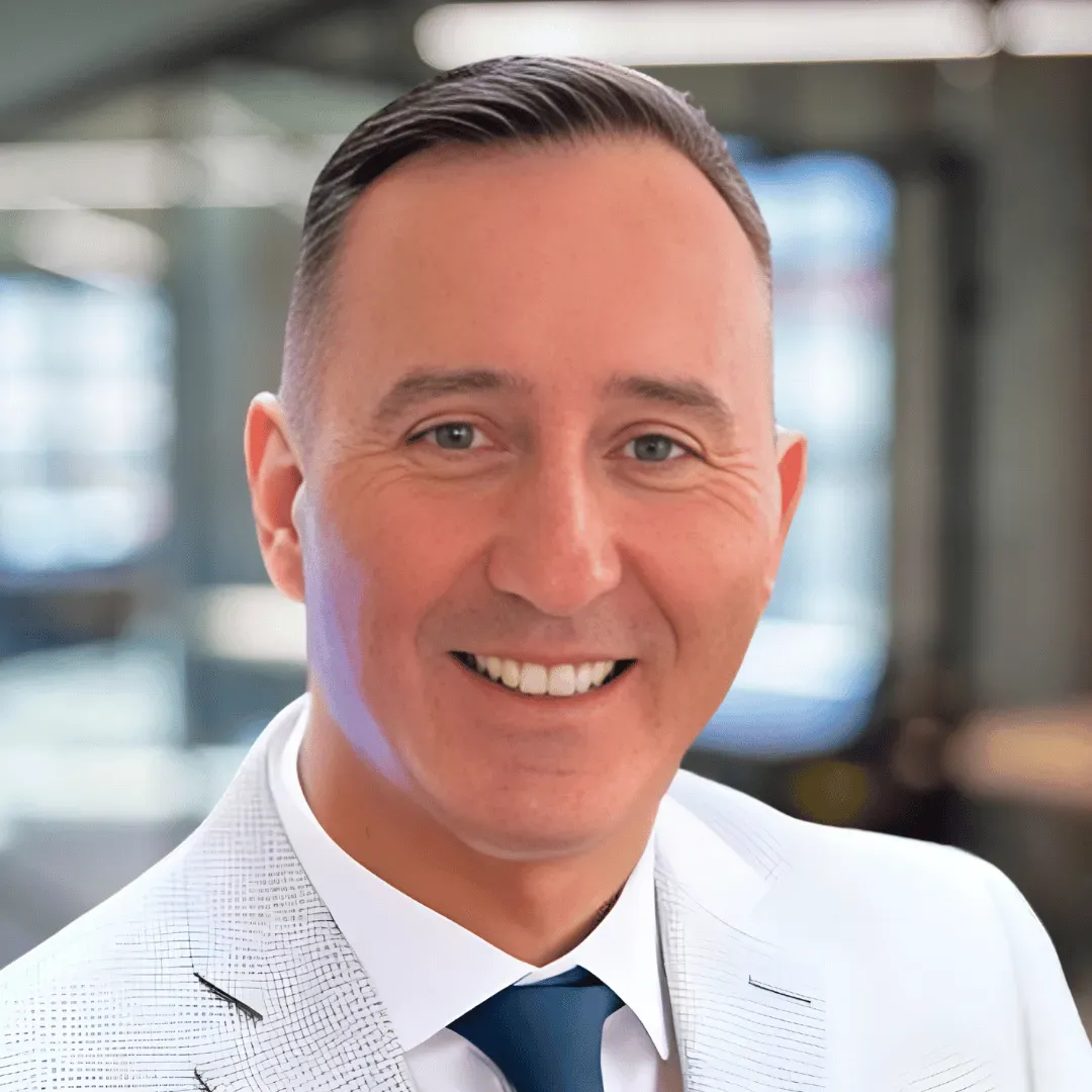 A tan man with black hair wearing a grey suite on a blurred background.