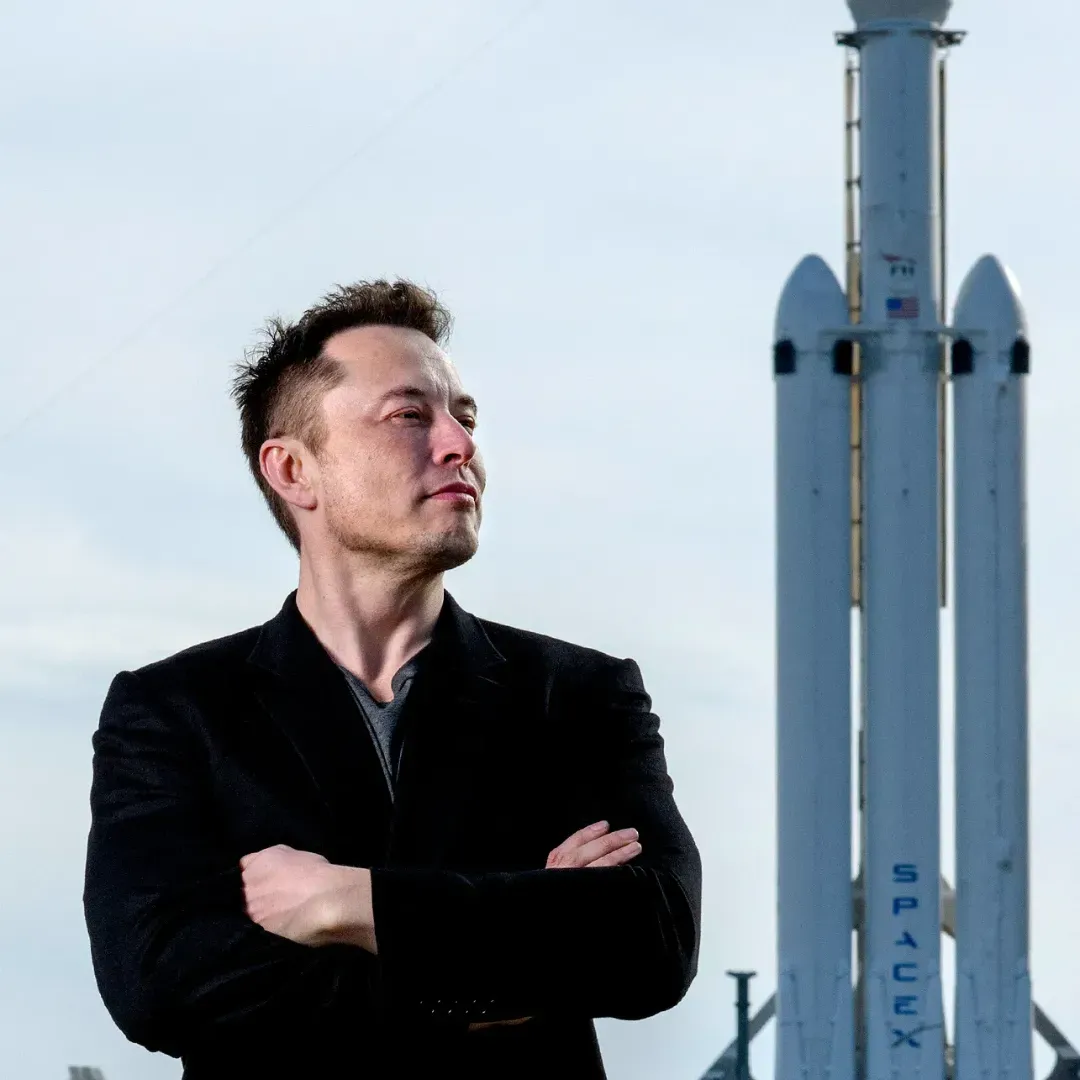 Elon Musk, CEO of Tesla and SpaceX with his arms crossed standing in front of a rocket ship.