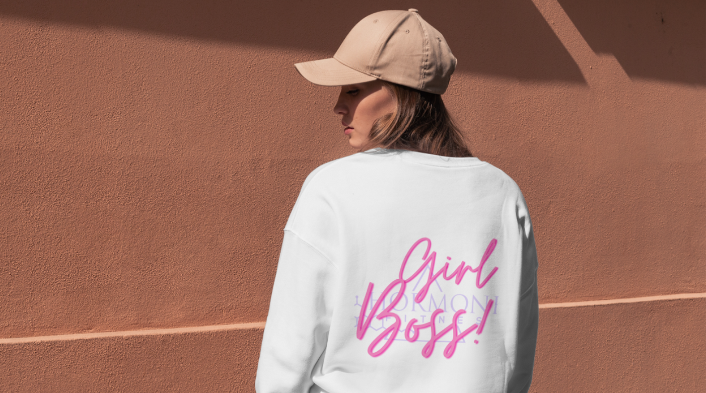 A woman wearing a white sweatshirt with a women empowerment quote on it standing in front of a wall.