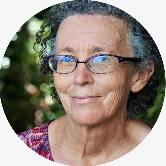 An headshot of an old woman with short grey and black hair wearing glasses is smiling 