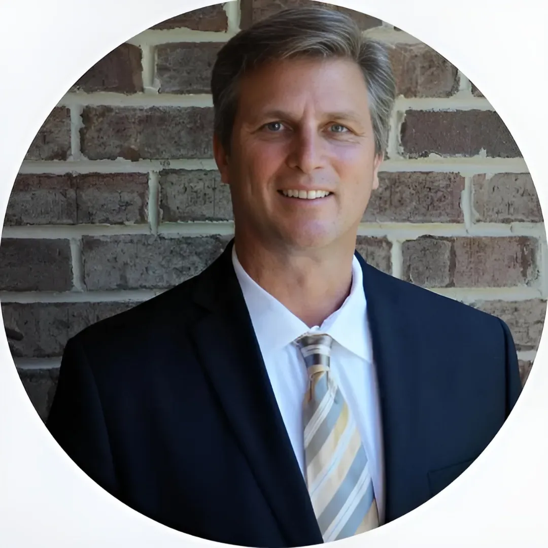 A headshot of a man in a black suit and multicolor tie stands in front of a brick wall