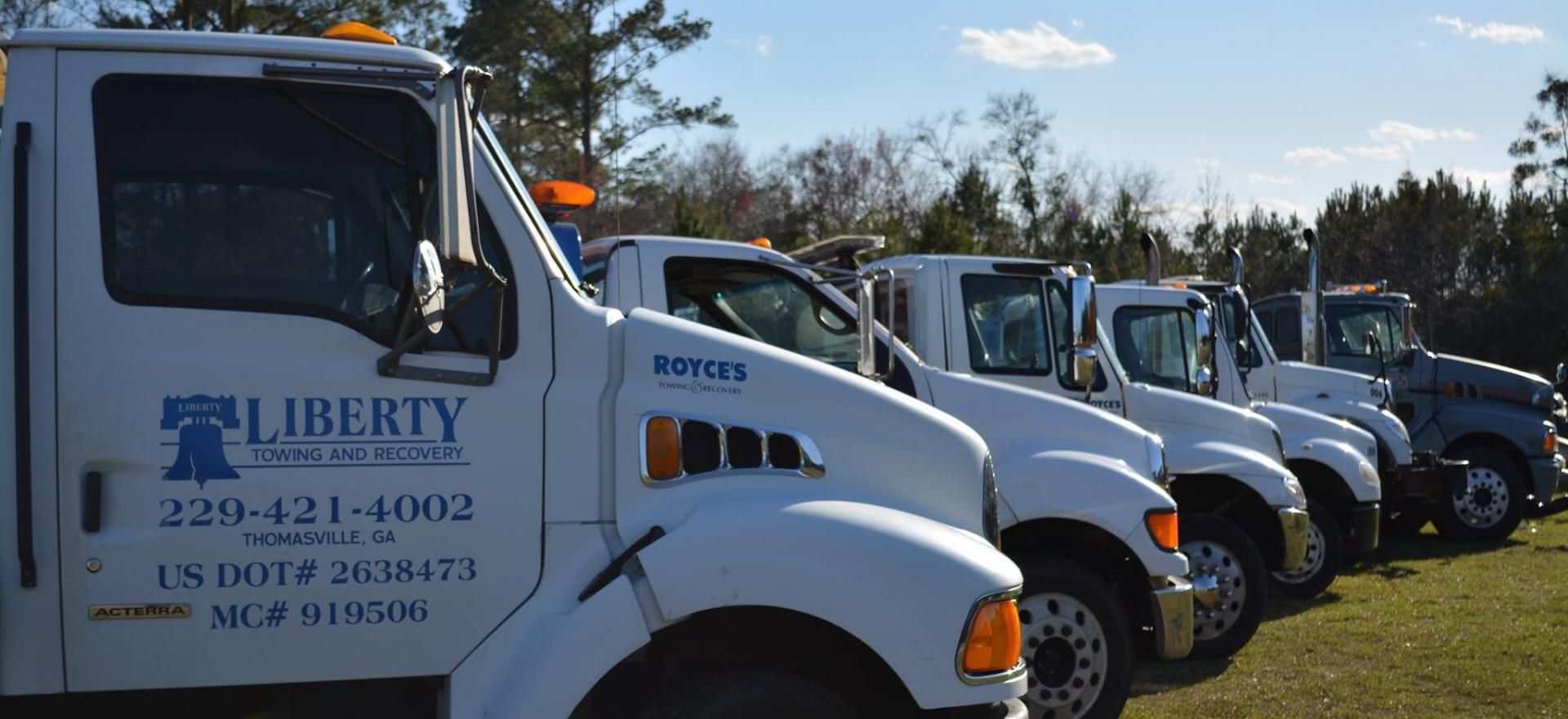 Row of white tow trucks parked on grass, under a blue sky. Liberty Towing logo on first truck.