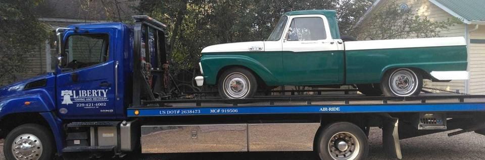 A green and white vintage pickup truck is on a blue tow truck, with a blue cab.