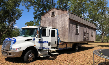 A large white truck carrying a small, weathered wooden barn on a sunny day.