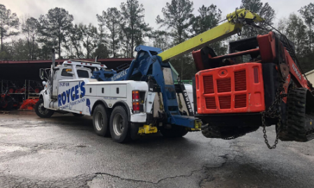 A tow truck lifting an orange skid steer loader in a lot. The truck is white with blue accents.