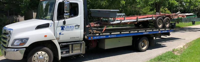 White flatbed tow truck hauling trailers on a sunny roadside.