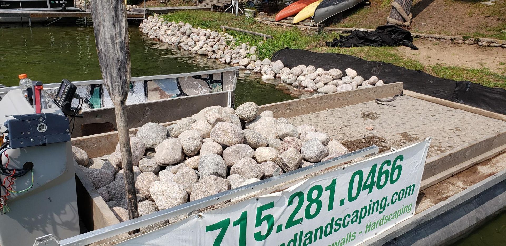 A boat filled with rocks near a shoreline;  banner displays a landscaping phone number.