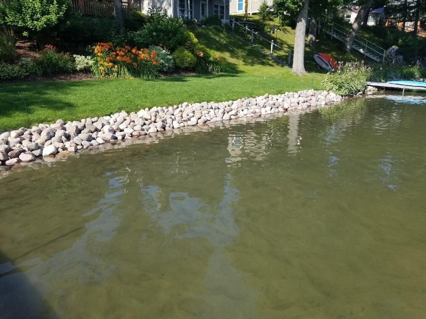 Lakeshore with a rock border, green grass, flowers, and water with reflections of trees and sky.