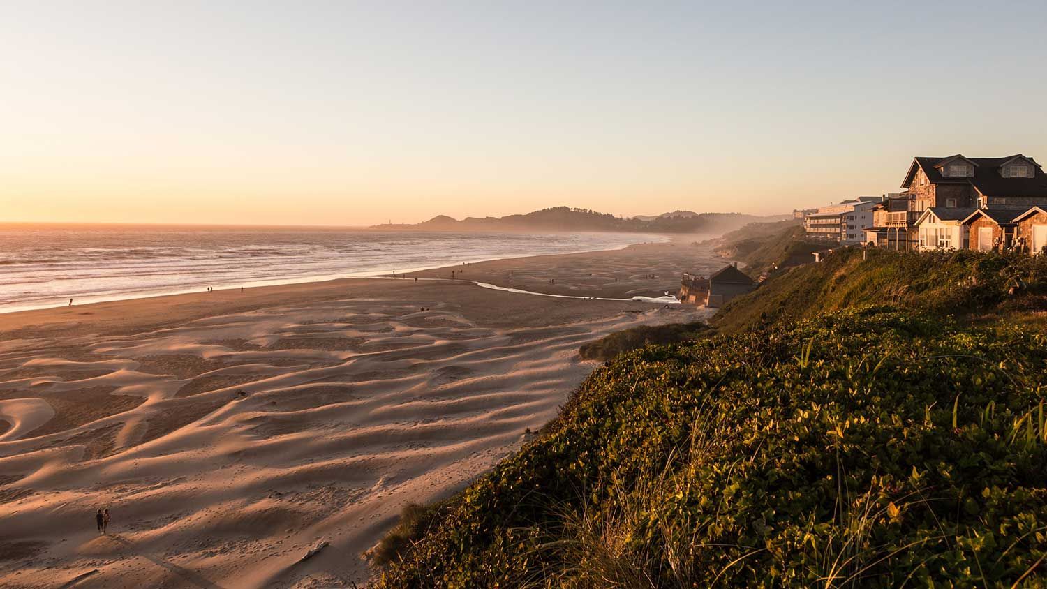 Sunset over a wide sandy beach with rolling waves and houses on a grassy bluff