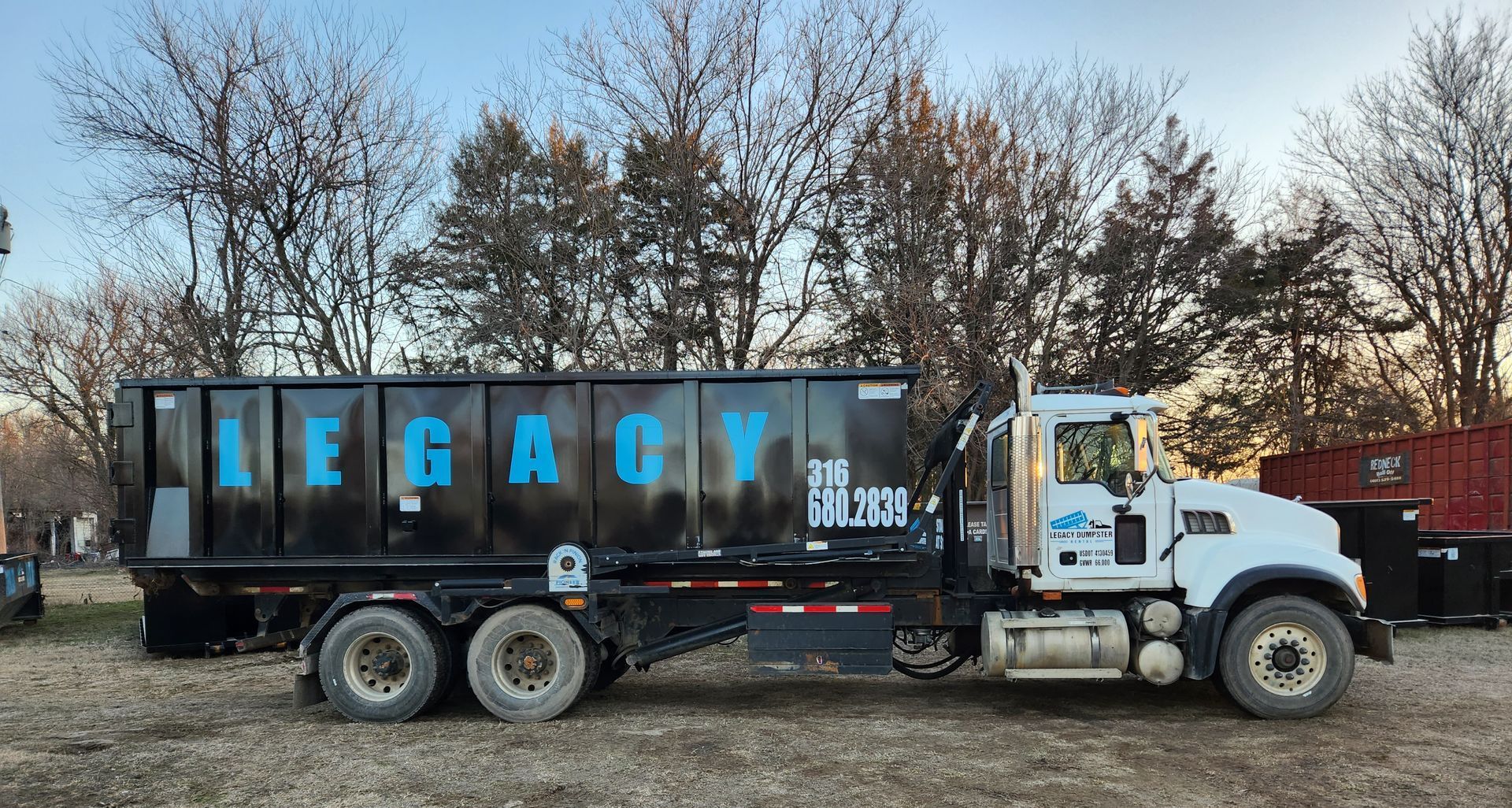 A dump truck is parked in a field with trees in the background and LEGACY with their business phone number on it