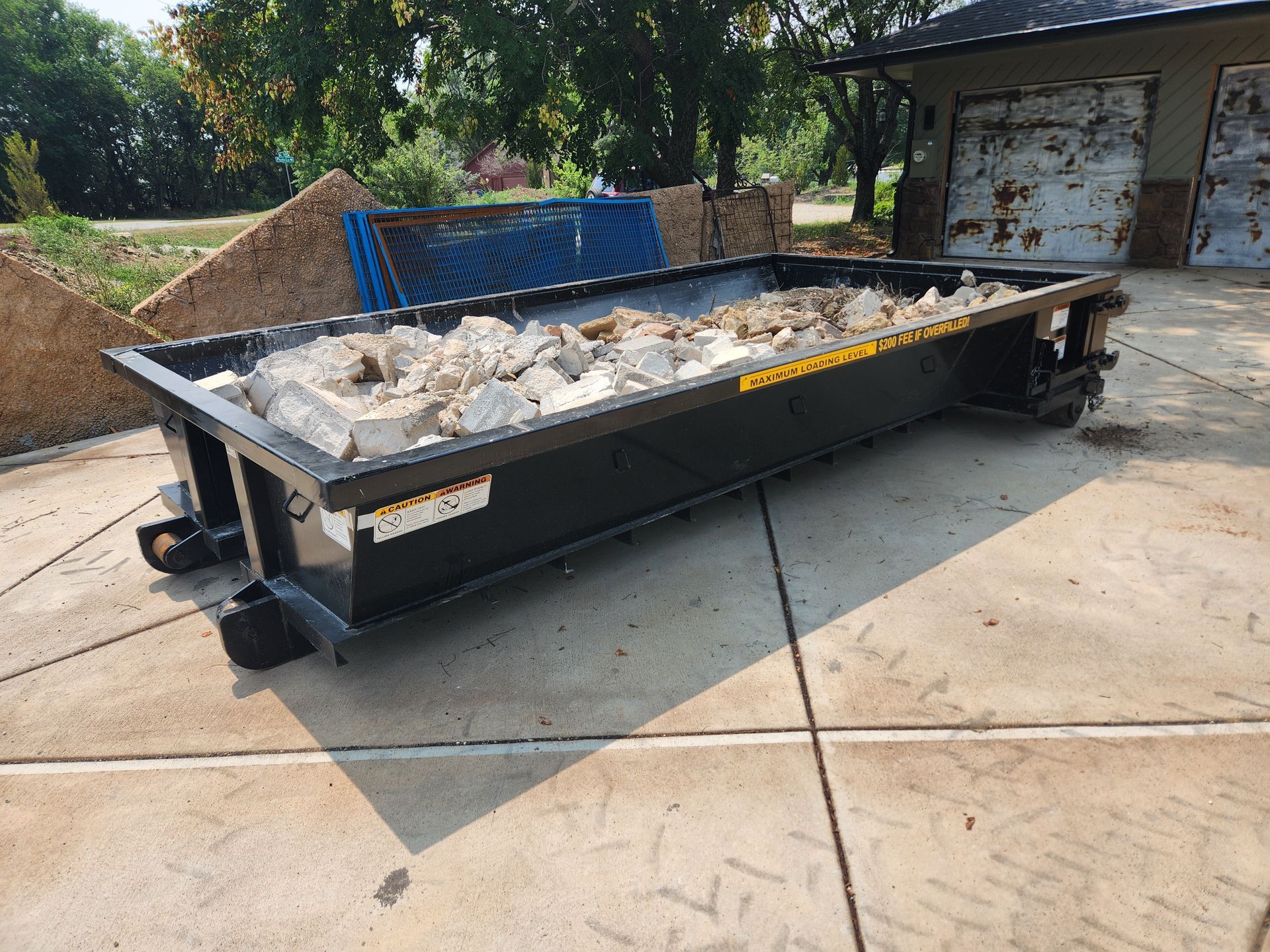 A dumpster filled with rocks is sitting on the sidewalk in front of a garage.