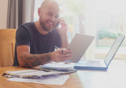 A man is sitting at a table with a laptop and a tablet while talking on a cell phone.