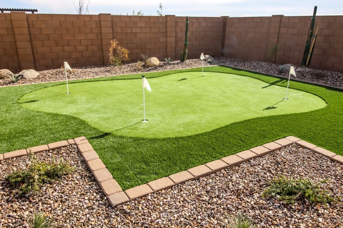 Backyard putting green with three holes and flags, surrounded by artificial turf, gravel, and a brick wall.