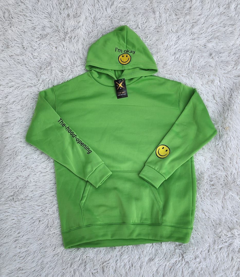 Una sudadera con capucha verde con una cara sonriente en las mangas está sentada sobre una alfombra blanca.