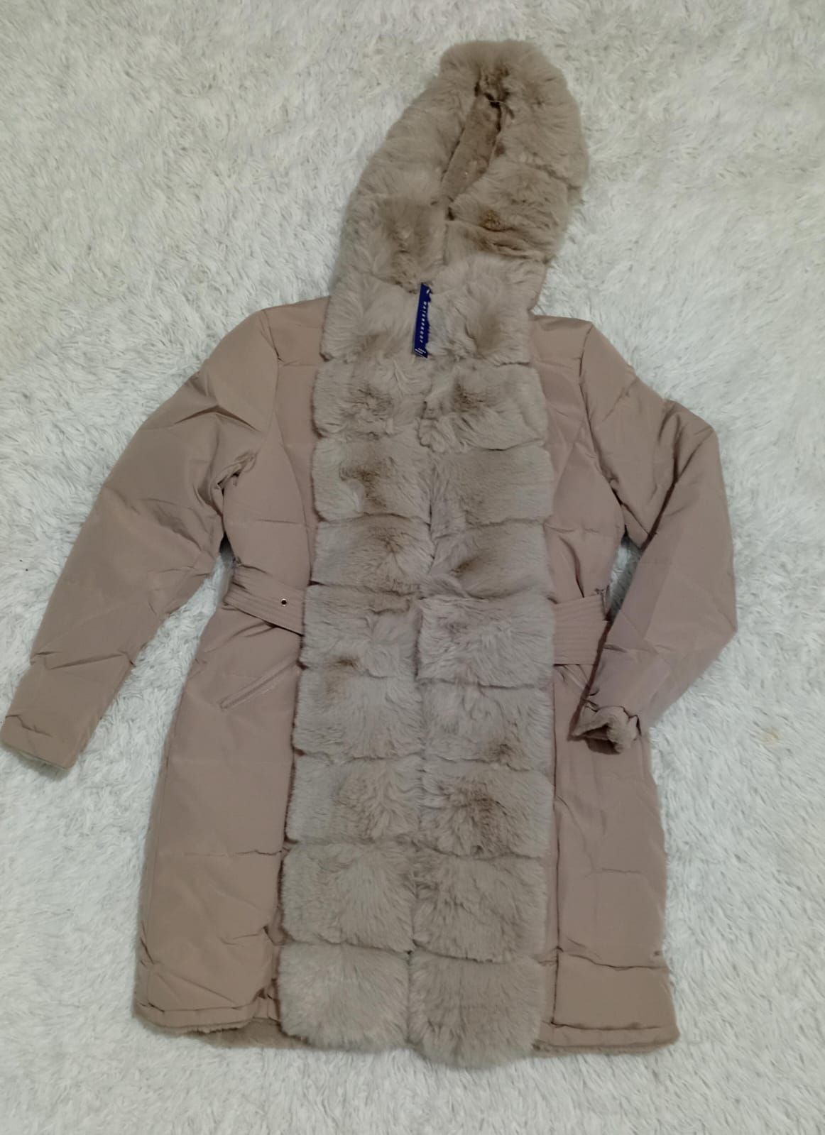 Un abrigo beige con capucha de piel está sentado sobre una alfombra blanca.