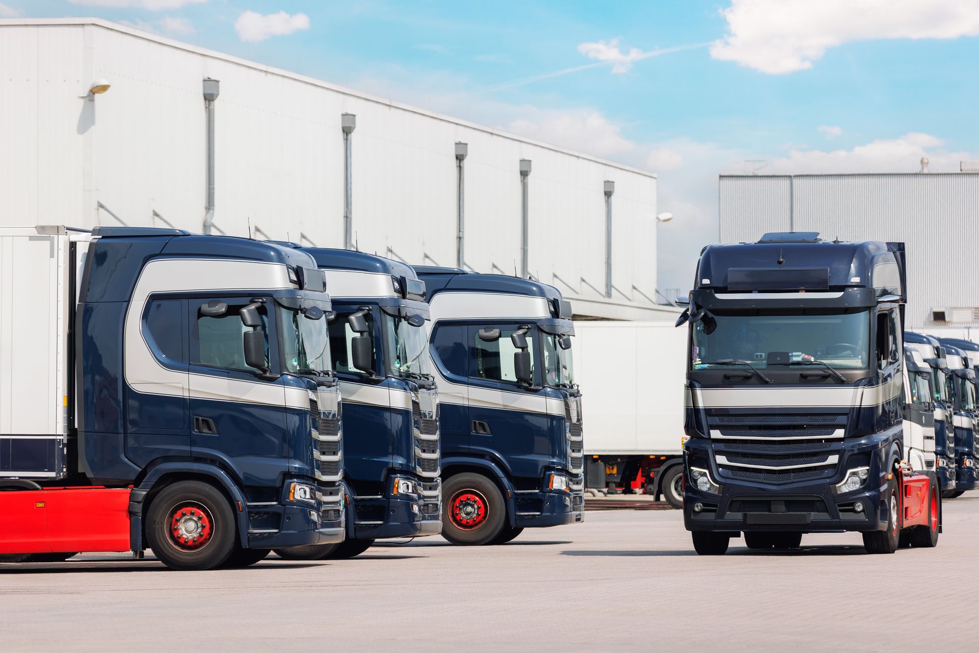 Blue and silver semi-trucks parked in a row in front of a warehouse on a sunny day.