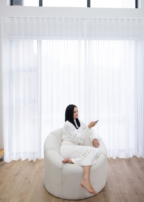 Woman relaxing in a white lounge chair in front of floor-to-ceiling lace curtains that brighten the minimalistic living space.