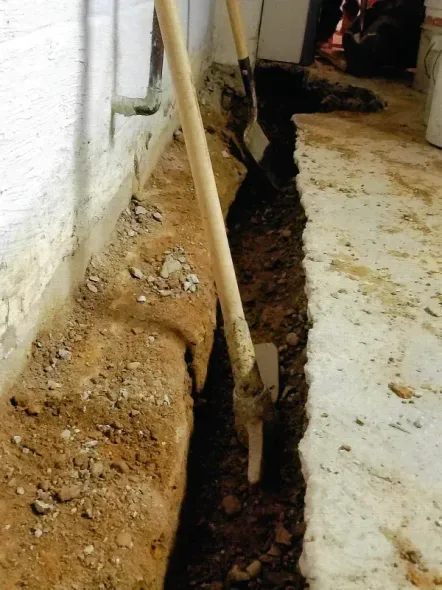 A trench dug along a basement wall beside a concrete floor, with a shovel standing inside the excavated soil and gravel.