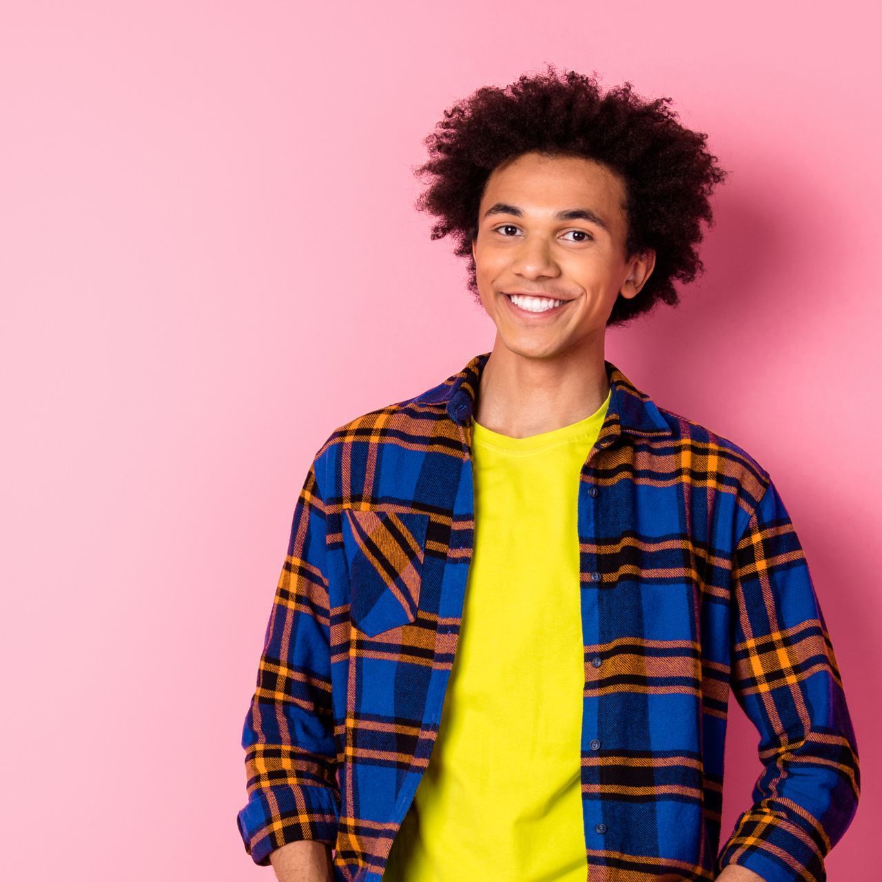 A young man wearing a plaid shirt and a yellow shirt is smiling against a pink background.