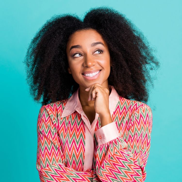 A woman with curly hair is smiling and looking up while holding her hand to her chin.