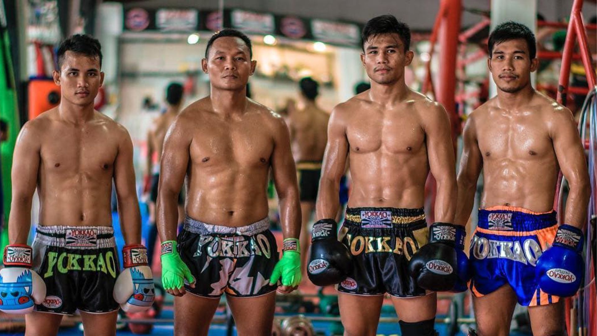 Four shirtless Muay Thai fighters wearing boxing gloves and shorts standing in a gym.