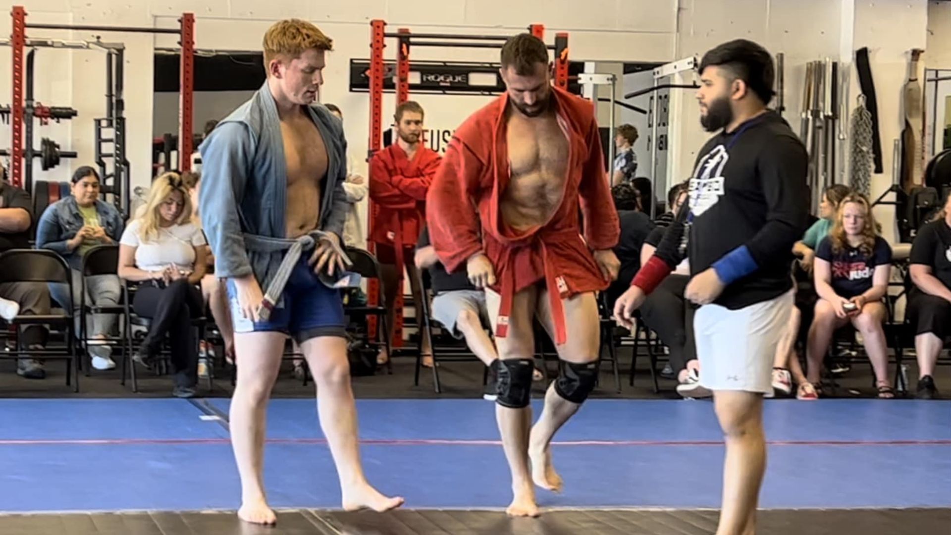 Athletes in sambo uniforms stand on a gym mat during a competition, with spectators seated in the background.