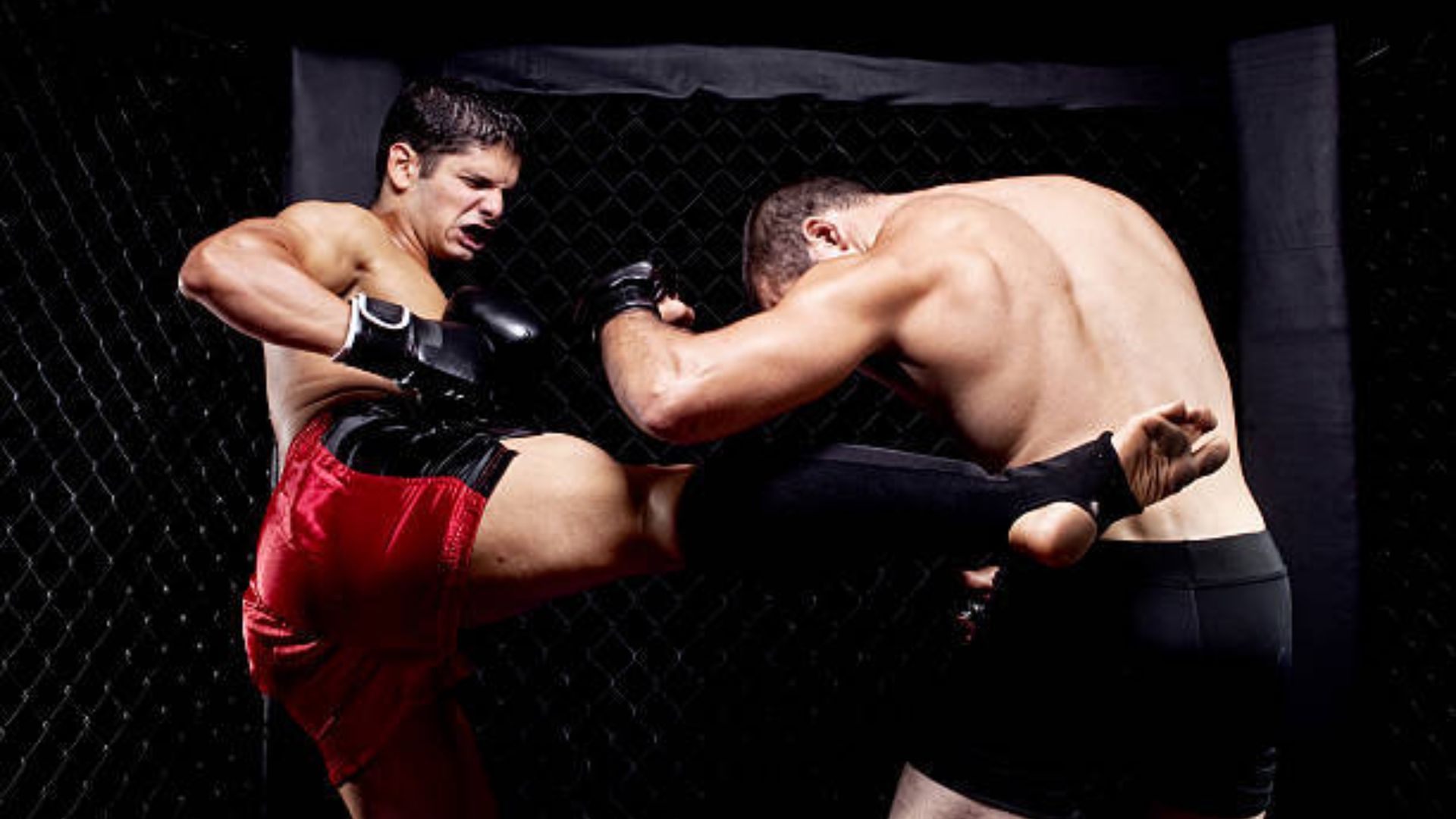 Two fighters in a MMA cage; one strikes with a kick to the side of the other, who wears black shorts and protective gear.