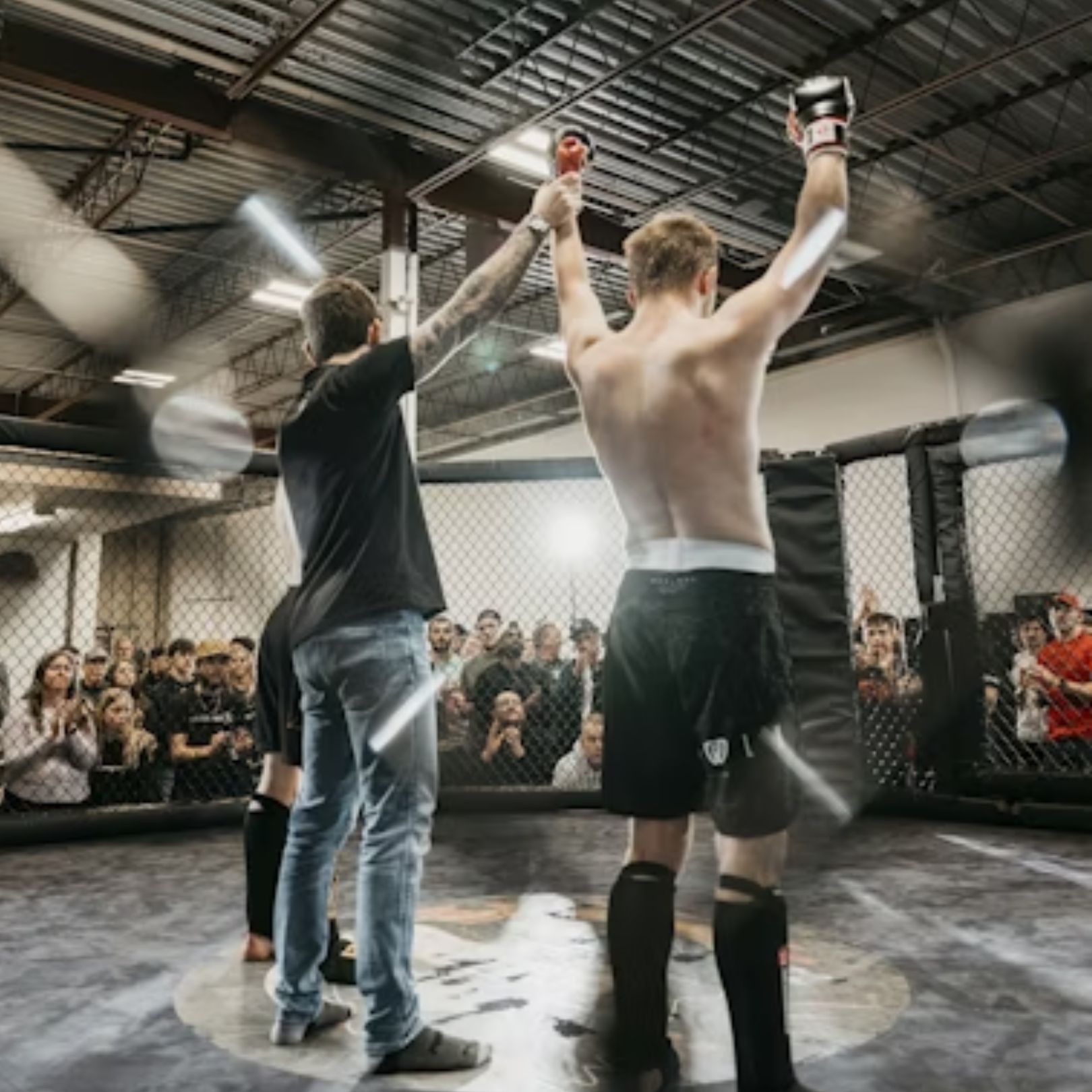 A referee raises a fighter's hand in victory inside an MMA cage, with spectators watching from the sidelines.
